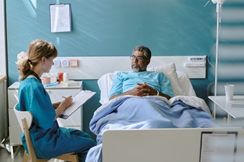 Caucasian female nurse sitting beside hospital bed interviewing middle aged biracial man lying under blanket, both engaged in conversation during medical consultation.