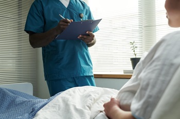 Black male nurse standing beside hospital bed holding clipboard, interacting with Caucasian pregnant female patient lying under blanket, medical consultation in progress.